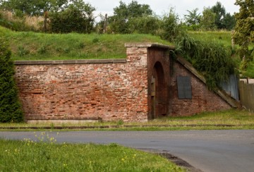 Zbytky strážnic brány nacházející se po levé straně, při pohledu do města. V popředí železniční koleje s pamětní deskou (na zdi), upomínající na osud obyvatel židovského ghetta.